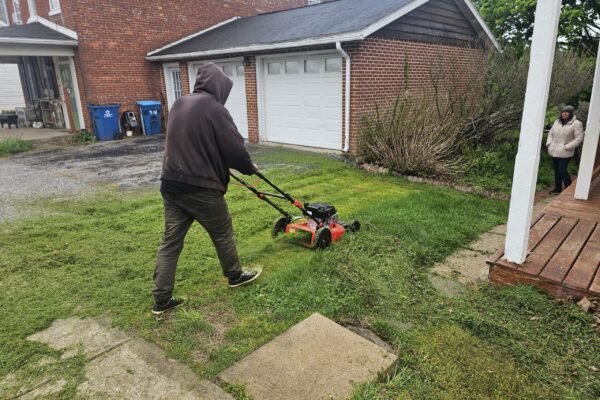 Person using a push mower to cut grass near a driveway and porch while another stands nearby