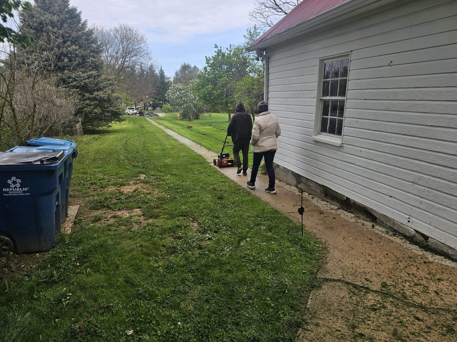 Person pushing a lawn mower along a narrow sidewalk path beside a house while another walks nearby