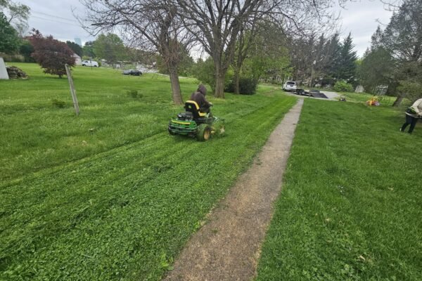 Riding mower cutting grass along a narrow path while another person trims the lawn nearby