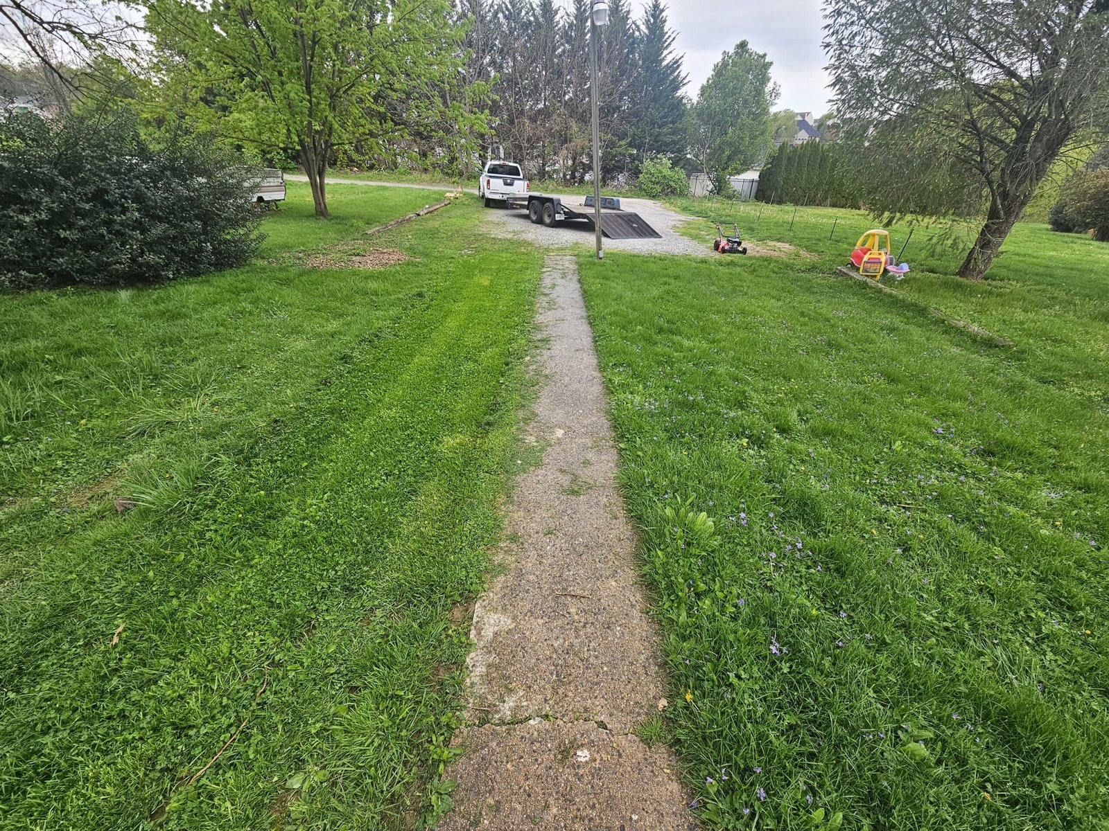 Narrow concrete walkway through a freshly cut lawn leading to a driveway with a trailer and mower in the background
